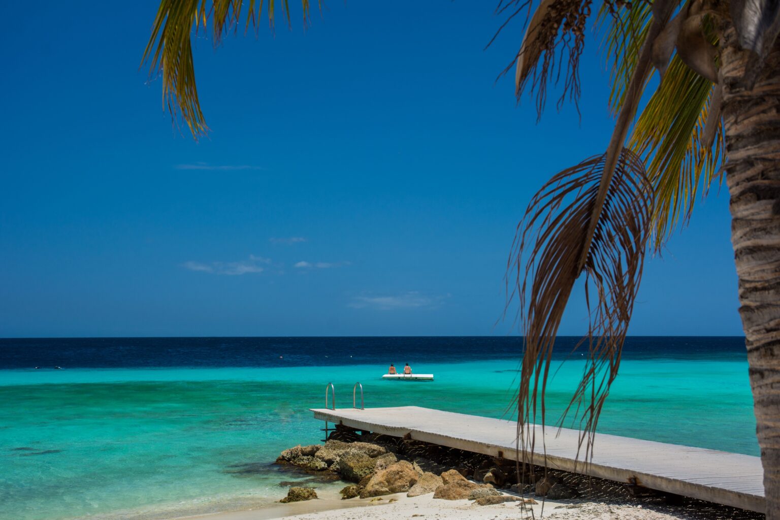 A tranquil beach scene with a wooden pier extending into turquoise water under a clear blue sky.